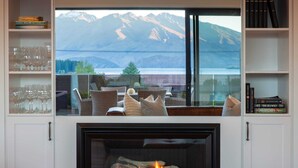 A fireplace with a mountain view, a bookshelf with books, and a glass display of wine glasses. - Waimaori Views The Sp (Wānaka)