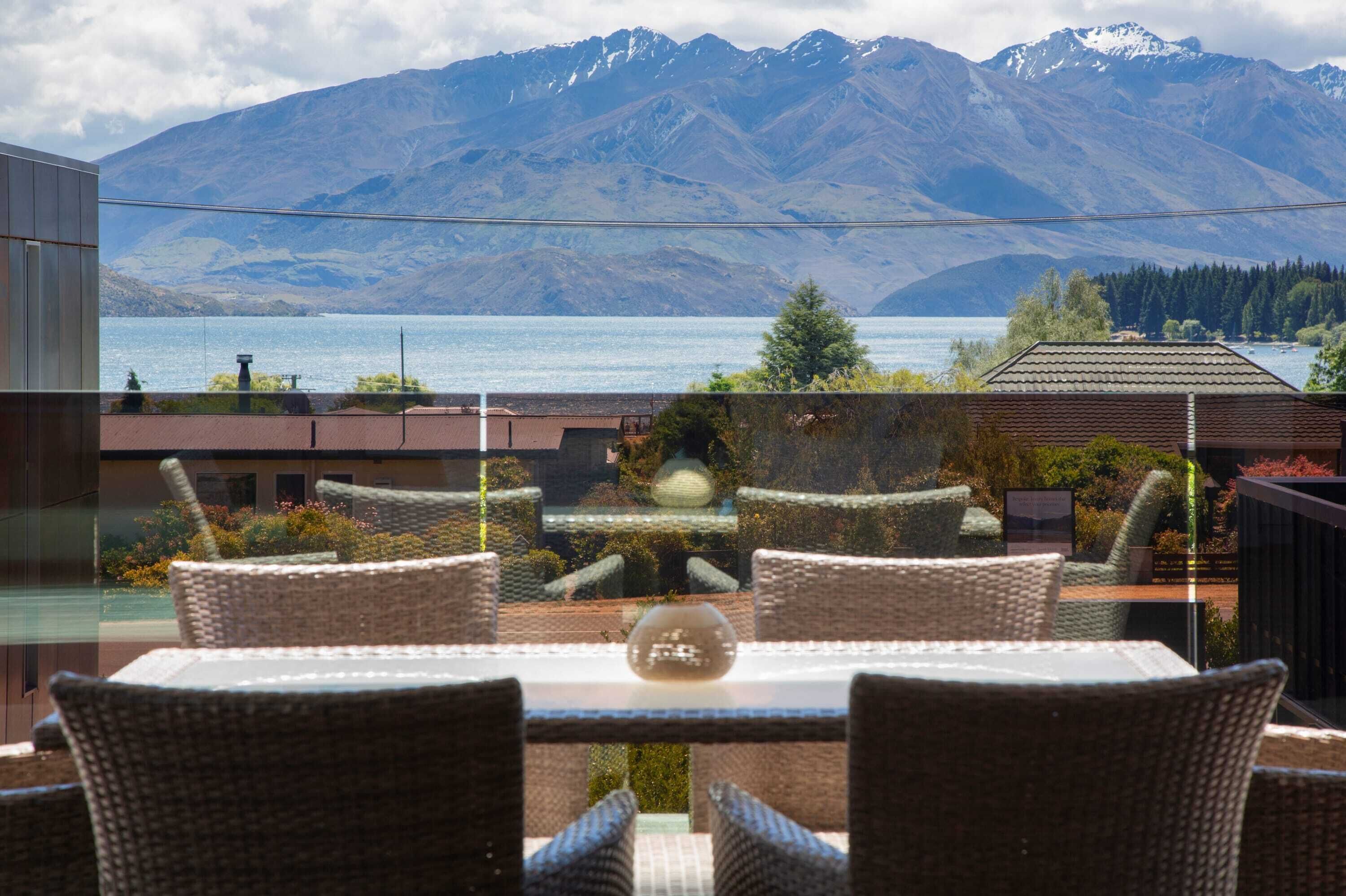 Una terraza con sillas de mimbre y una mesa, con vistas a un lago y montañas.