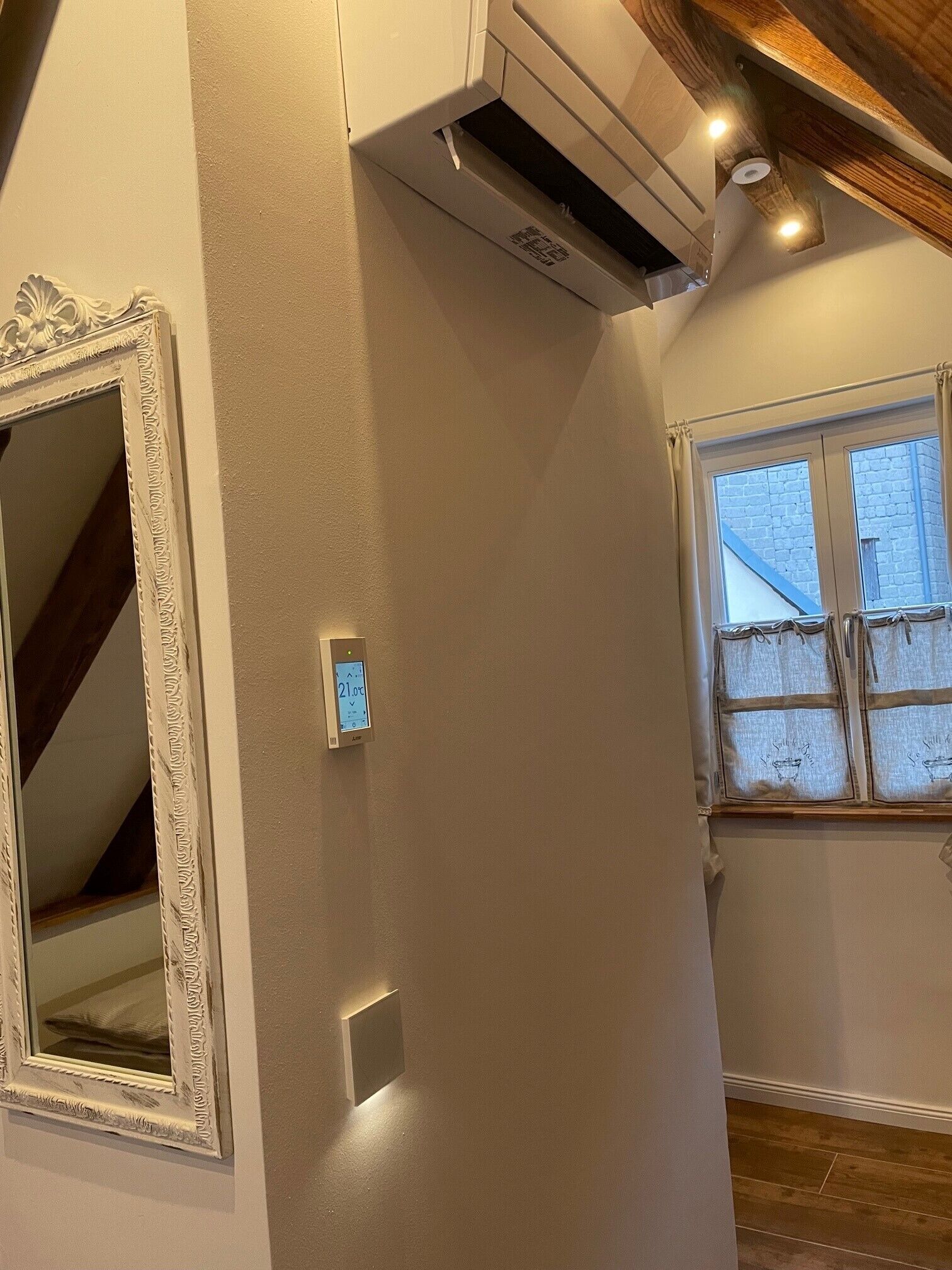 Una habitación con un espejo grande, un equipo de aire acondicionado, un termostato y una ventana con vista a un edificio de ladrillo.