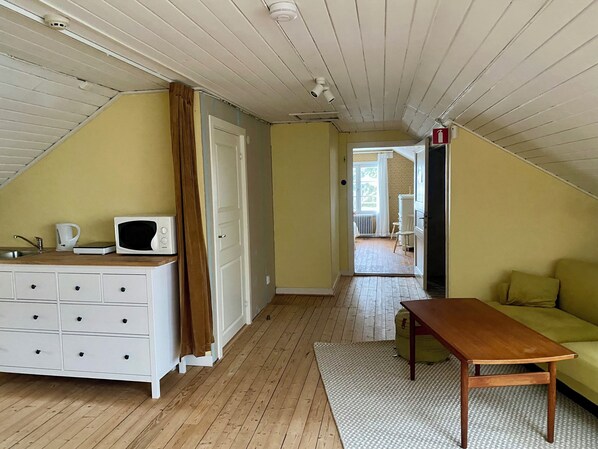 A room with a sloped ceiling, a white dresser with drawers, a microwave, a wooden table, and a green sofa. - Lappnäs Bed & Breakfast (Sunne)