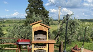 Un espacio al aire libre con cocina de ladrillo, que incluye una parrilla y un horno de leña, rodeado de follaje y un paisaje pintoresco.