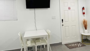 Una habitación con una mesa y sillas blancas, un televisor de pantalla plana montado en la pared, una puerta blanca con un extintor y una pequeña mesita de noche con un florero.