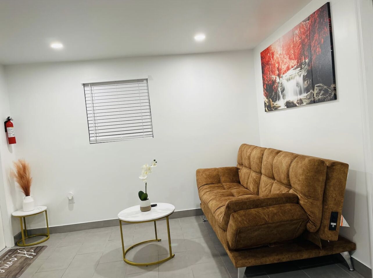 A living room with a brown sofa, a white round coffee table, a small white side table with a vase and plant, a window with blinds, and a painting on the wall.
