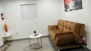 Un salón con un sofá marrón, una mesa de centro redonda blanca, una pequeña mesita auxiliar blanca con un jarrón y una planta, una ventana con persianas y un cuadro en la pared.