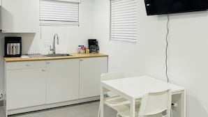 Una cocina pequeña y moderna con armarios blancos, encimera de madera y cafetera integrada. Hay una mesa de comedor blanca con cuatro sillas y un televisor de pantalla plana montado en la pared.