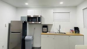 Una cocina compacta con armarios blancos, un refrigerador de acero inoxidable, un microondas, una cafetera y un lavamanos.