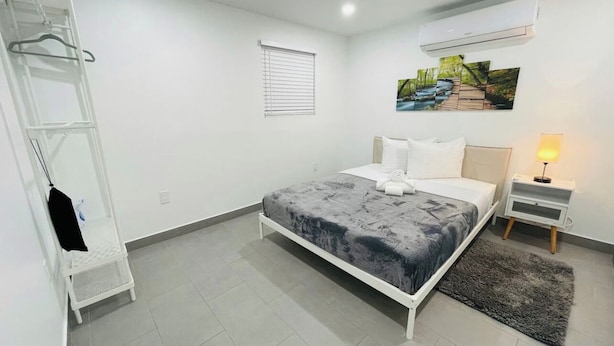 Un dormitorio con cama, mesita de noche con lámpara, aire acondicionado instalado en la pared, ventana con persianas y armario blanco con una prenda colgada.