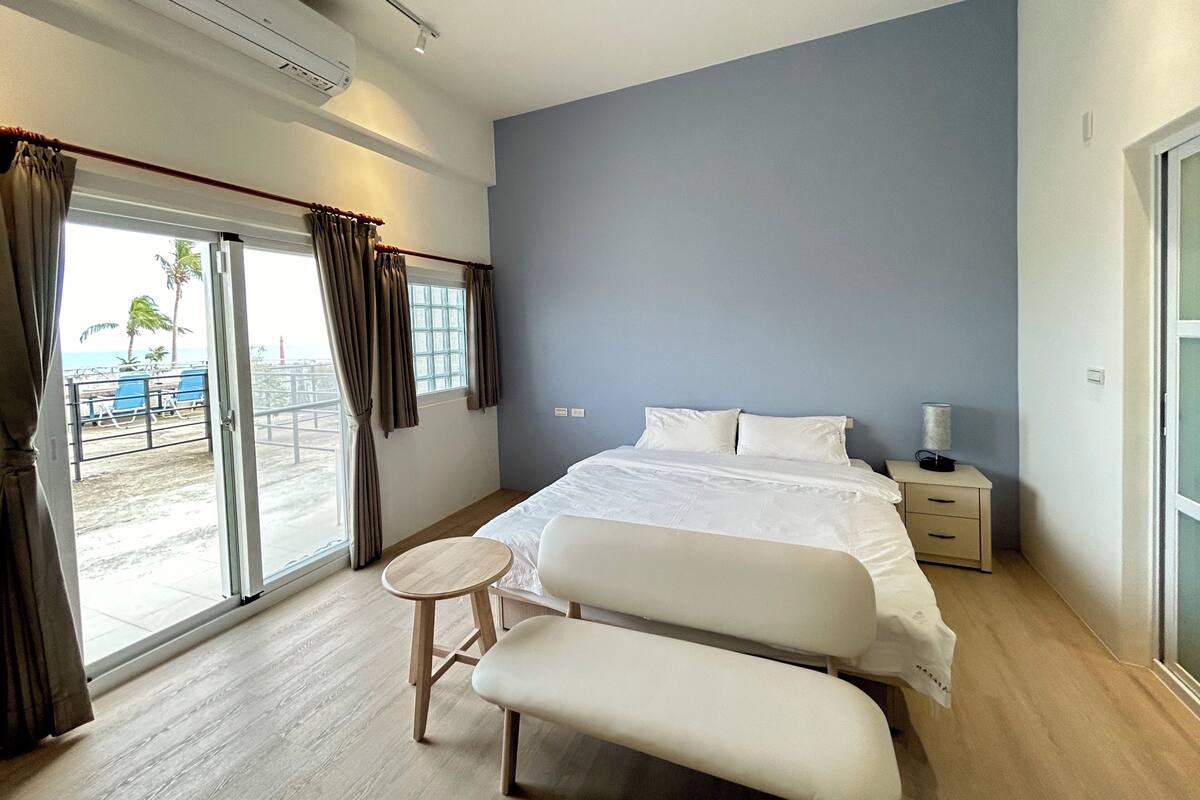A modern hotel room with a large bed, a bedside table with a lamp, a small round table, and a bench. The room has a sliding glass door leading to a balcony with a view of the beach and palm trees.