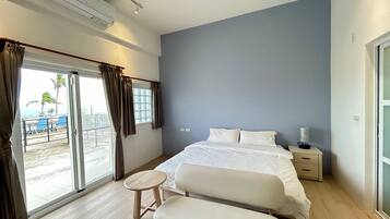 A modern hotel room with a large bed, a bedside table with a lamp, a small round table, and a bench. The room has a sliding glass door leading to a balcony with a view of the beach and palm trees.