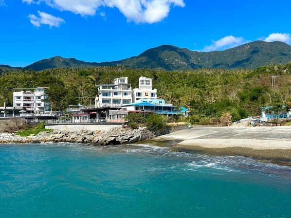 Um hotel à beira-mar com vários edifícios, uma praia e montanhas ao fundo.