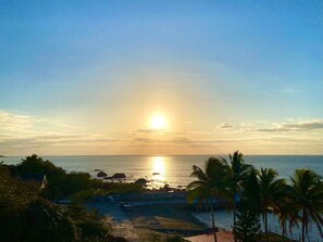 Vista costeira com um pôr do sol, palmeiras e uma estrada que leva a uma praia.