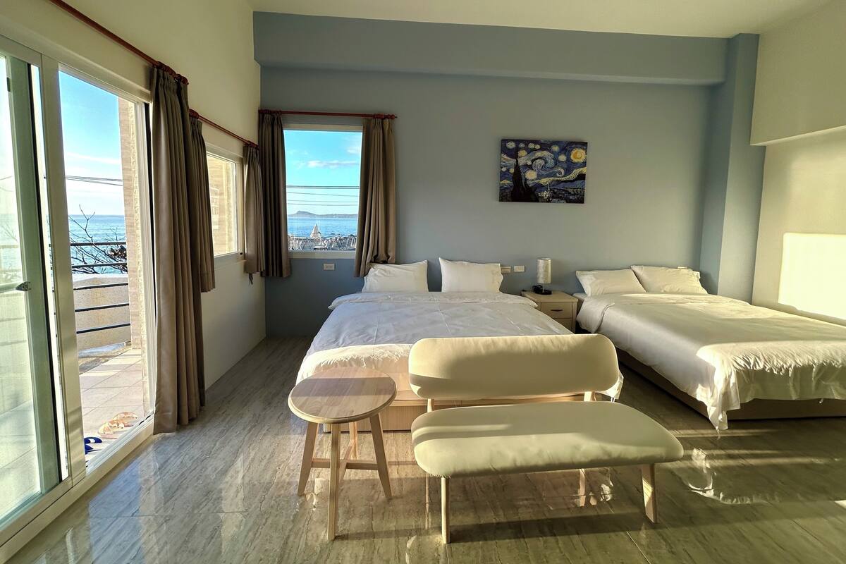 A hotel room with two beds, a small table, a bench, and a painting on the wall.