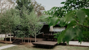 Una terraza de madera con una mesa y sillas, rodeada de árboles y un edificio con zona cubierta.