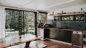 A modern kitchen with a dining table, built-in appliances, and large windows overlooking a garden. - The Artemis Cottage - Romantic Couples Retreat (Woodlands)