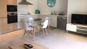 A modern kitchen with built-in appliances, a dining area with a glass table and white chairs, and a flat-screen TV on a wooden stand.