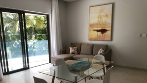 A modern living room with a glass dining table, white chairs, a beige sofa, and a large window with a view of greenery.