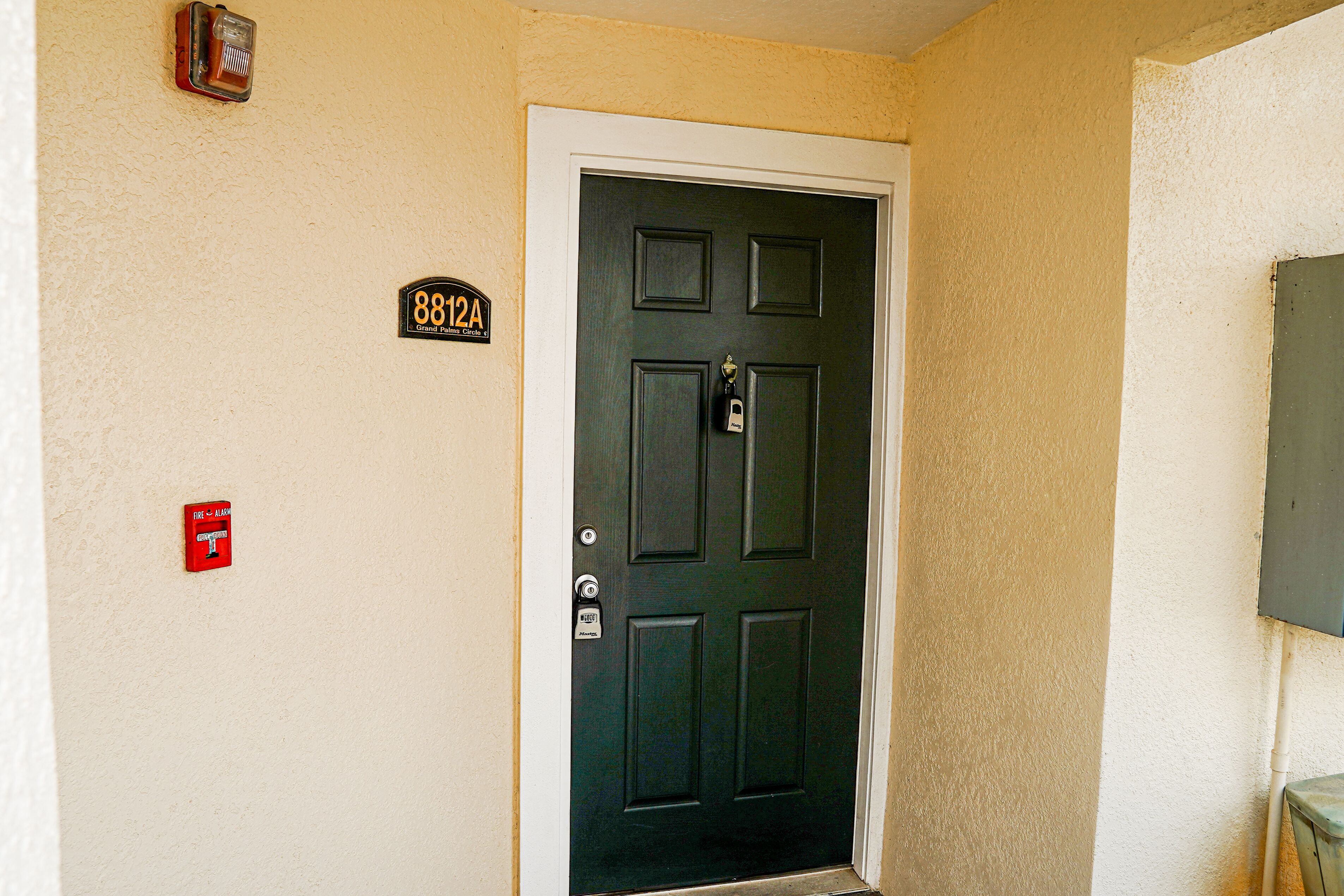 A black door with a keypad lock, a fire alarm, and a house number sign.