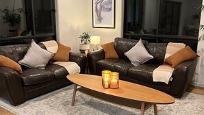 A living room with two leather sofas, a wooden coffee table, and a black leather ottoman. There is a ceiling fan, a framed picture on the wall, and a window with a view.