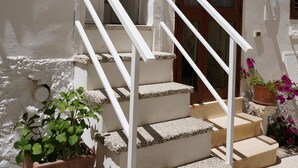 Un edificio blanco con puerta de madera, escaleras blancas con barandilla de metal y macetas con plantas.