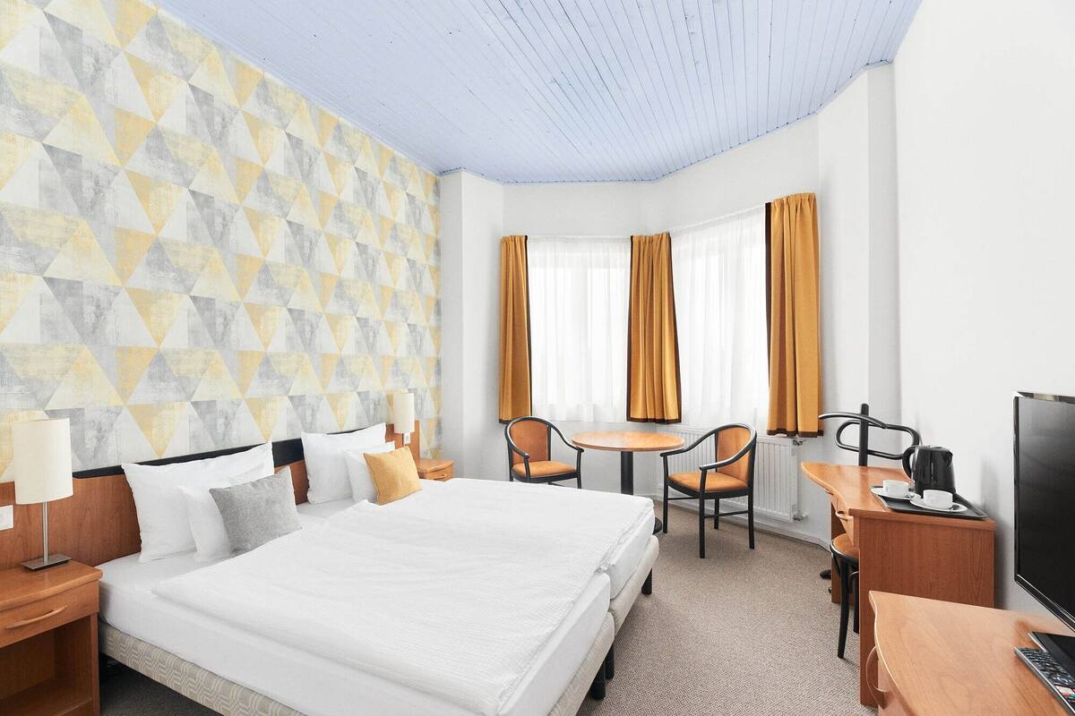 A hotel room with a large bed, a desk with a TV, a chair, a small table, and a window with curtains.