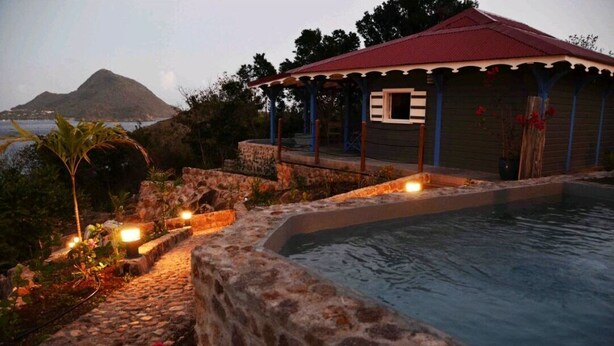 A poolside area with a stone wall, a covered seating area, and a view of a mountain and water. - Cases créoles classified 4**** with private pools (Terre-de-Bas)