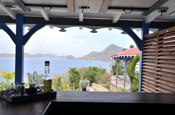 A bar area with a view of the sea and mountains, featuring a bottle of rum, glasses, and a tray.
