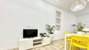 A modern living room with a flat-screen TV on a white entertainment unit, a dining table with yellow chairs, and a hanging light fixture. - Mejorana Apartament (1 bedroom apartment) (Madrid)
