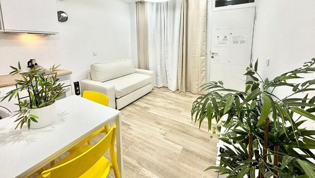 A modern living room with a white sofa, a dining table with yellow chairs, and a kitchen area with a white countertop. - Mejorana Apartament (1 bedroom apartment) (Madrid)