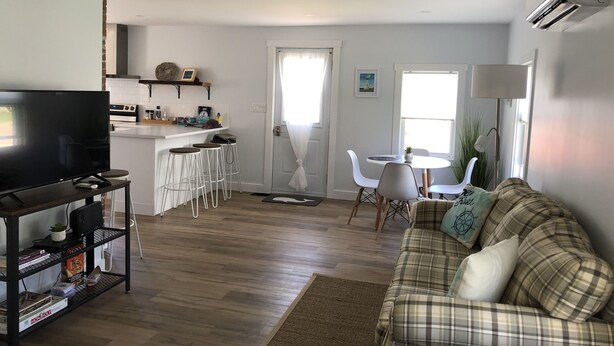 A modern living room with a plaid sofa, a TV on a stand, and a kitchen area with a bar. - Peaceful vacation home located in the heart of St Andrews-by-the-Sea (St. Andrews)