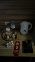 Una mesa de madera con una botella de agua, cafetera, bolsita de té y diversos bocadillos. Se observan dos controles remotos y un cartel que dice 'No molestar'.
