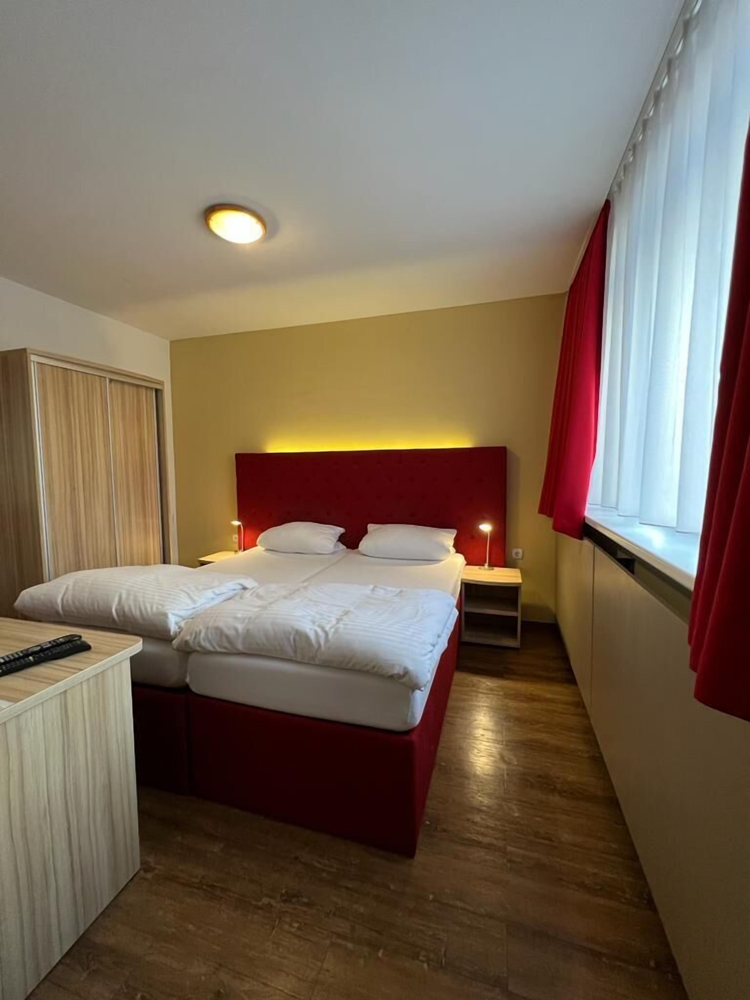 Une chambre d’hôtel avec un lit double, une table de chevet, un bureau avec une chaise, une armoire et une fenêtre avec des rideaux.