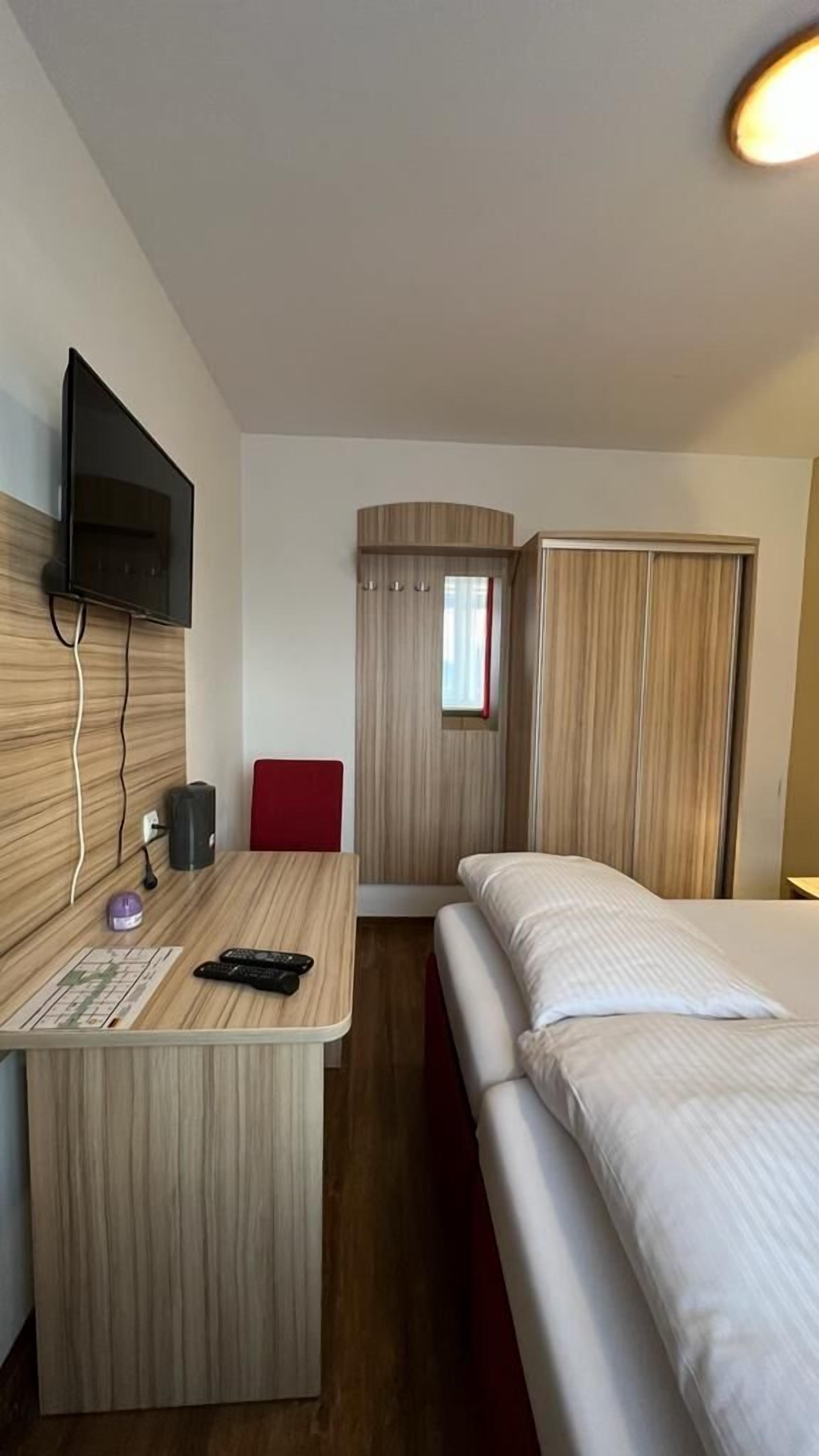 Quarto de hotel com cama, escrivaninha com cadeira, televisão fixada na parede e janela com cortinas.