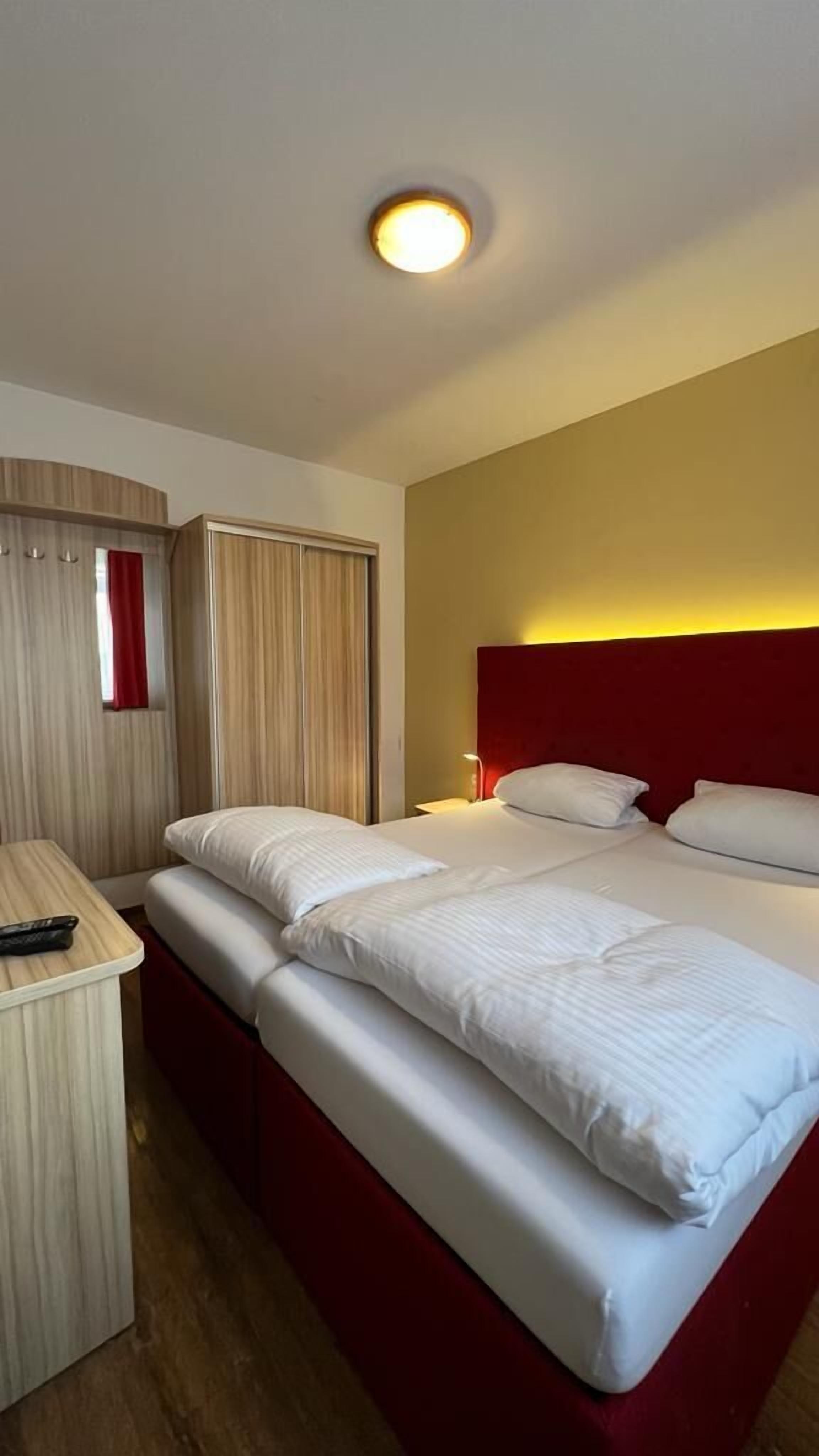 Ein Hotelzimmer mit einem großen Bett, einem Nachttisch, einem roten Kopfteil und einer Deckenleuchte.