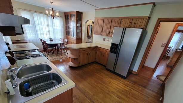Een keuken met houten kastjes, een roestvrijstalen koelkast, een eethoek met stoelen en een raam met gordijnen.