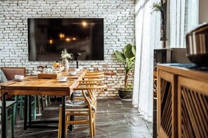 Een eetruimte met een lange houten tafel, rieten stoelen en een witte bakstenen muur.