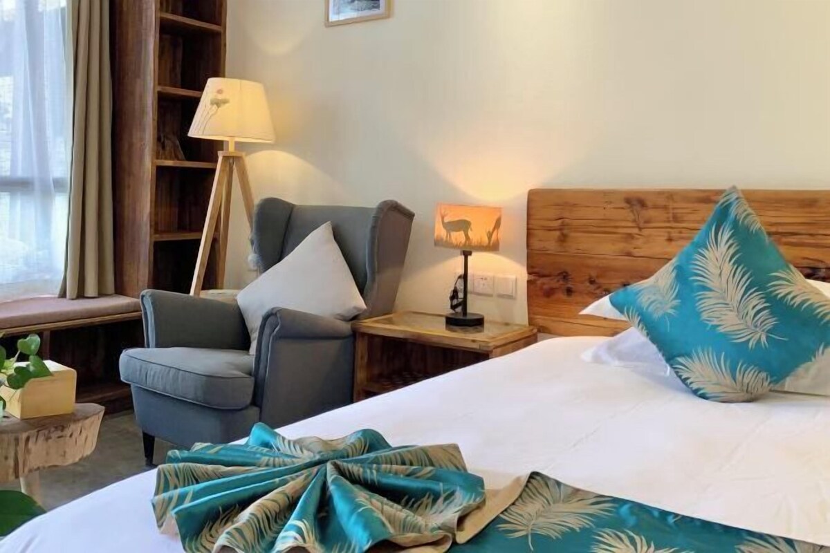 A hotel room with a bed, a chair, a lamp, a nightstand, a wooden headboard, and a window with curtains.