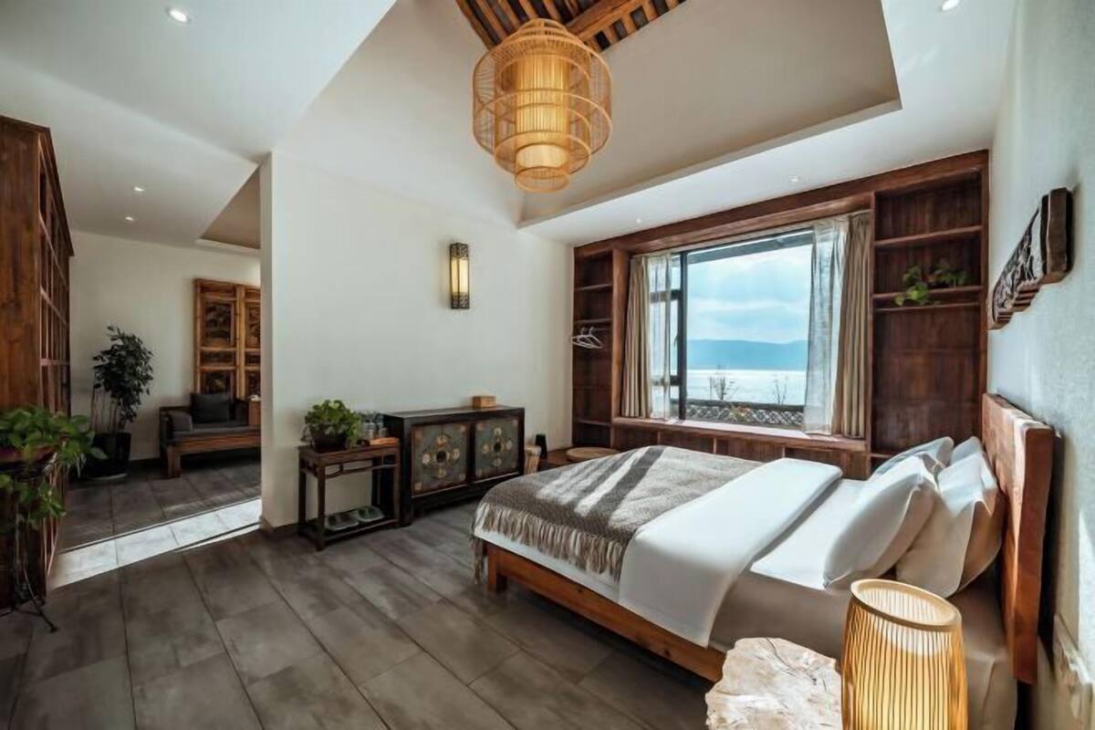 A bedroom with a large bed, wooden headboard, a window with a view, a bedside table, and a hanging light fixture.