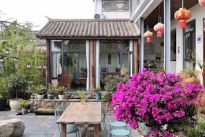 Een traditioneel gebouw in Chinese stijl, met een betegeld dak, een houten tafel met stoelen, en felroze bloemen op de voorgrond.