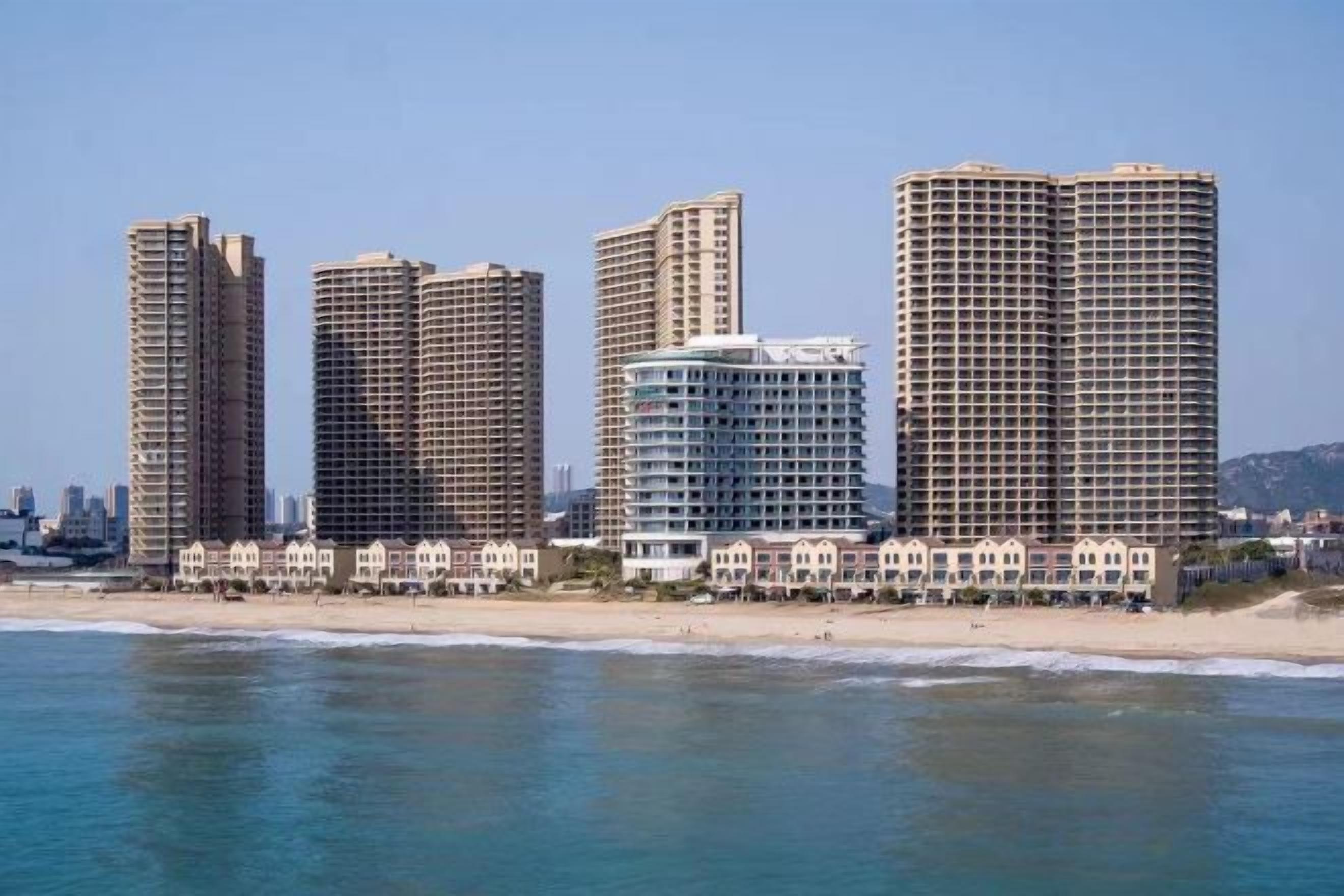 Un hotel frente a la playa con varios edificios altos, una playa de arena y un cielo azul despejado.