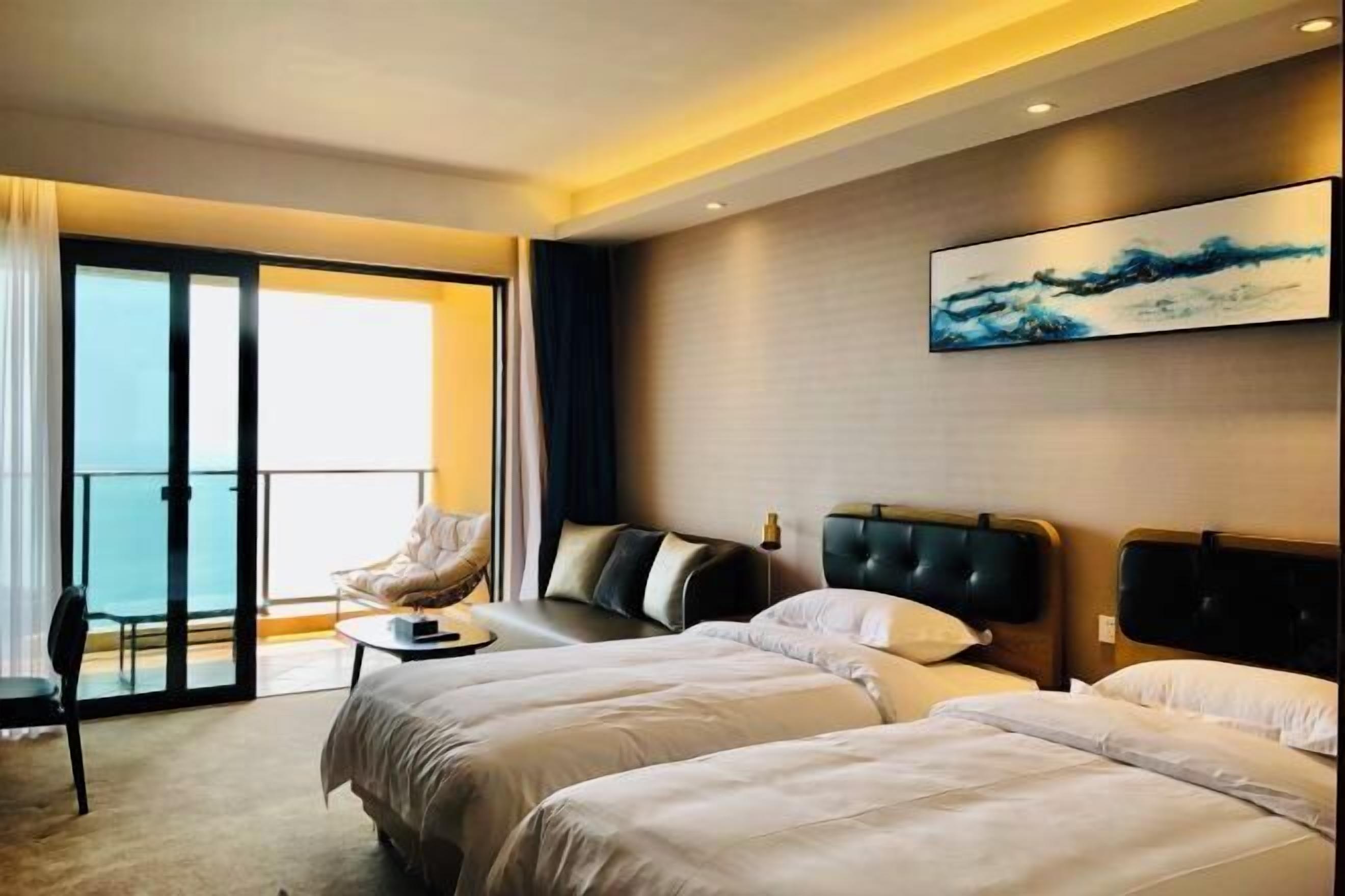 Quarto de hotel com duas camas, uma cadeira, uma mesinha e uma janela grande com vista.