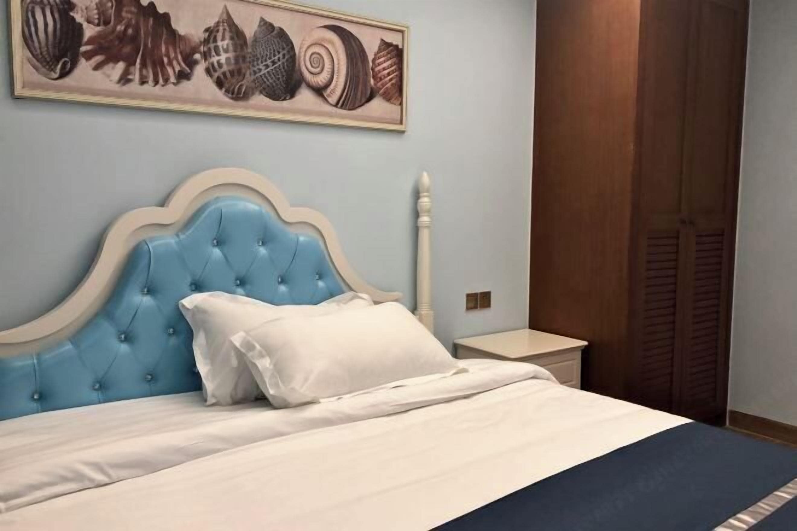 Un dormitorio con cabecera azul, colcha blanca y un cuadro de conchas marinas colgado en la pared.