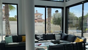 Un salon moderne avec un canapé d’angle, une table basse et un coin repas avec des chaises. De grandes fenêtres offrent une vue sur les palmiers et les bâtiments environnants.