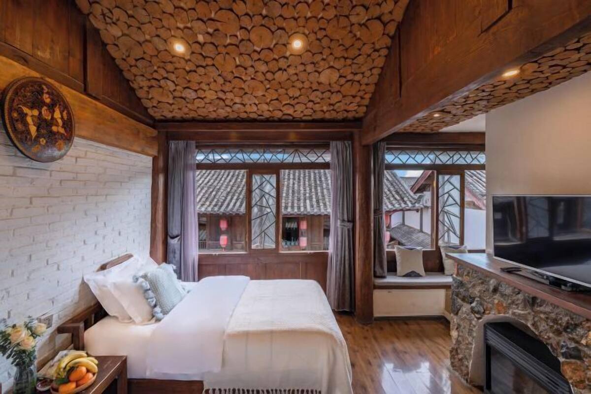 A bedroom with a bed, a TV, a stone fireplace, and a window with a view of traditional buildings.