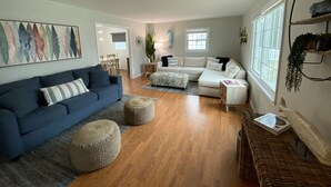 A spacious living room with a blue sofa, a white sectional sofa, and a coffee table. There is a large window providing natural light and a view of the outdoors. - Bayside Retreat: Your Perfect Beach Getaway (Lower Township)