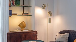 A cozy living room with a wooden cabinet, a small round table, a blue glass table, a sofa with cushions, a floor lamp, and shelves with books and decorative items. - Le Cementine in Lecce (Lecce)