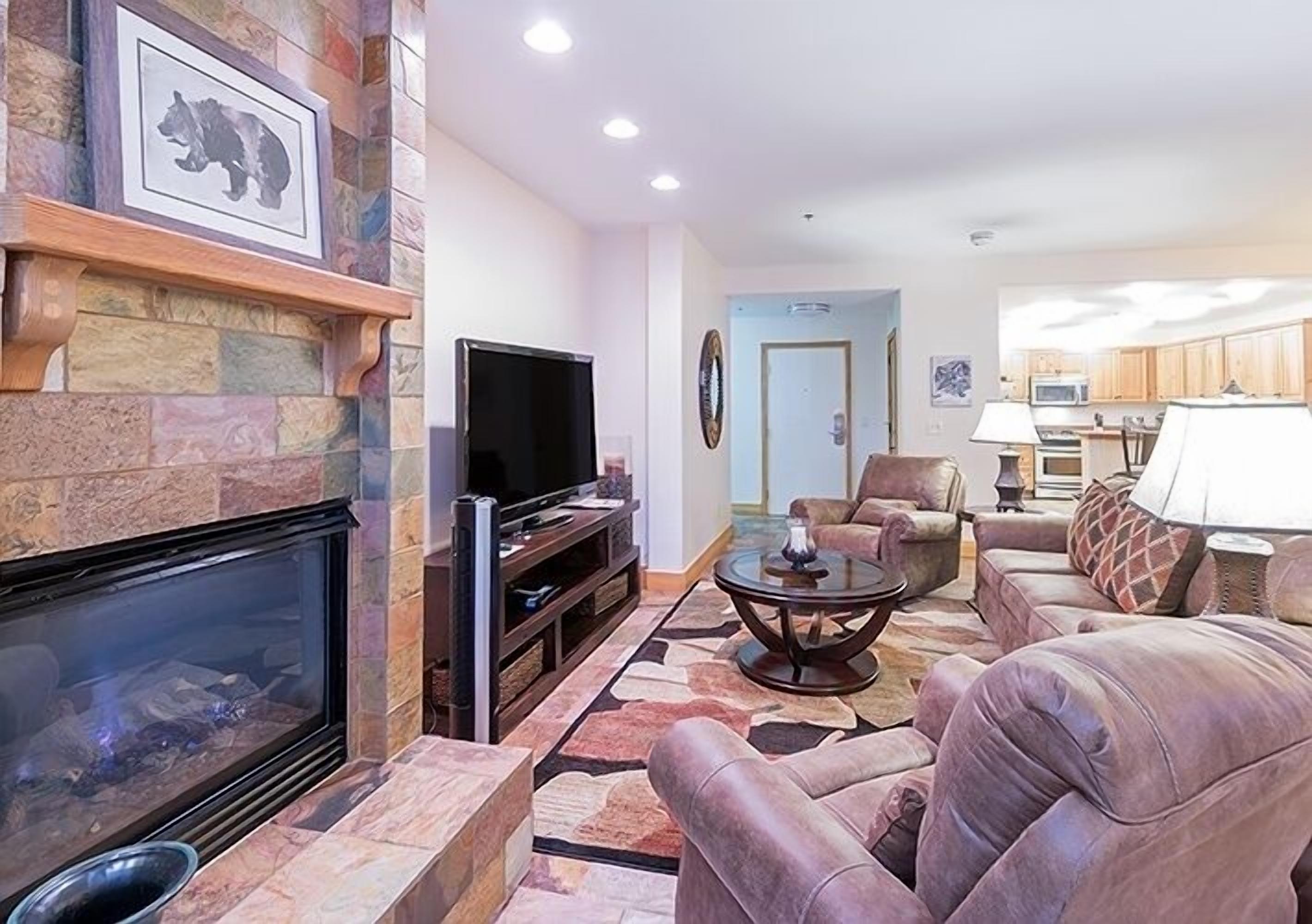 Una sala de estar con chimenea de piedra, sofás de cuero, un televisor de pantalla plana y una cocina al fondo.