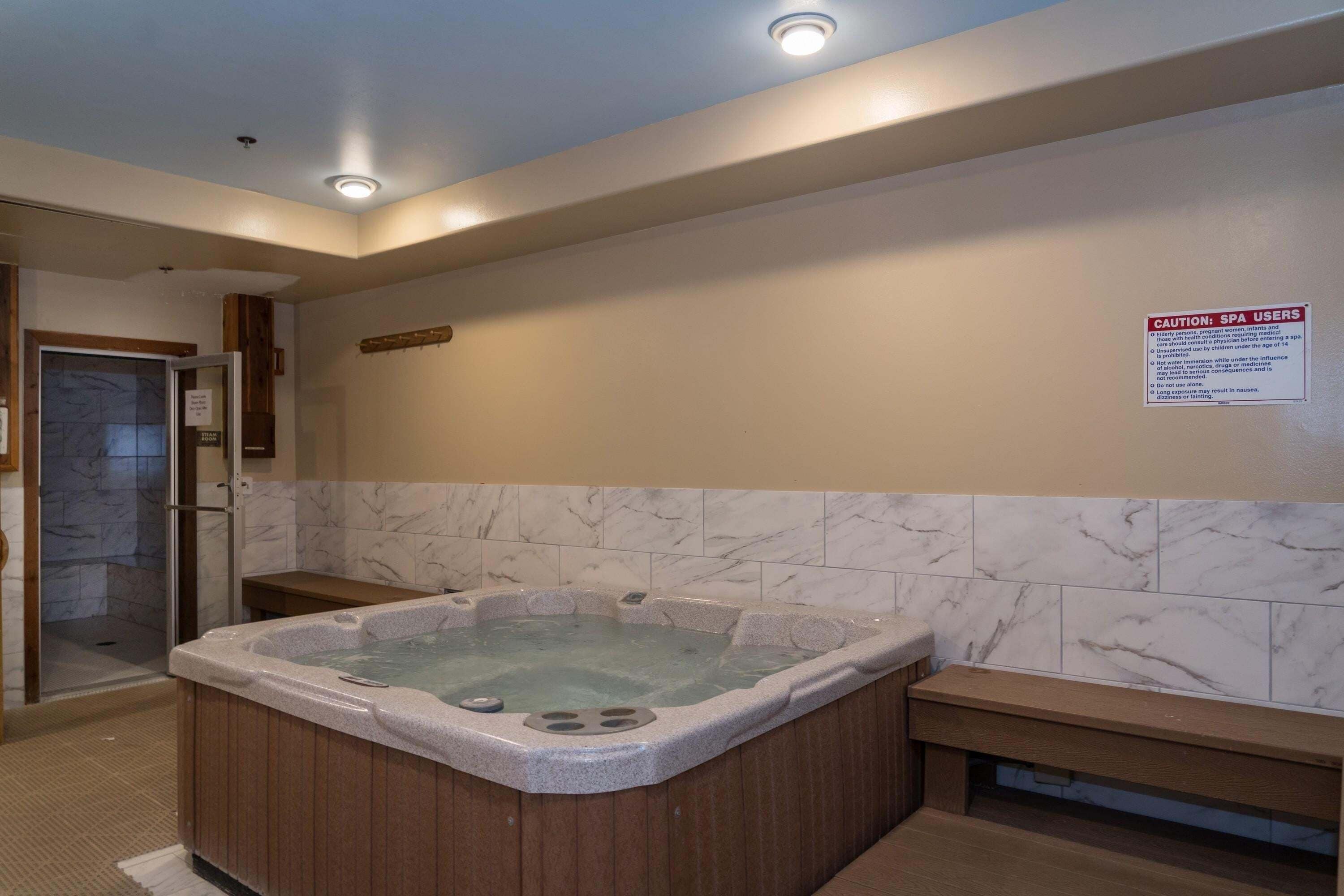 A spa area with a hot tub, wooden benches, a glass shower enclosure, and a caution sign for spa users.