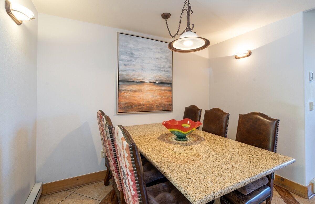 Een eetruimte met een granieten tafel, zes stoelen, een decoratieve schaal en een schilderij van een zonsondergang aan de muur.