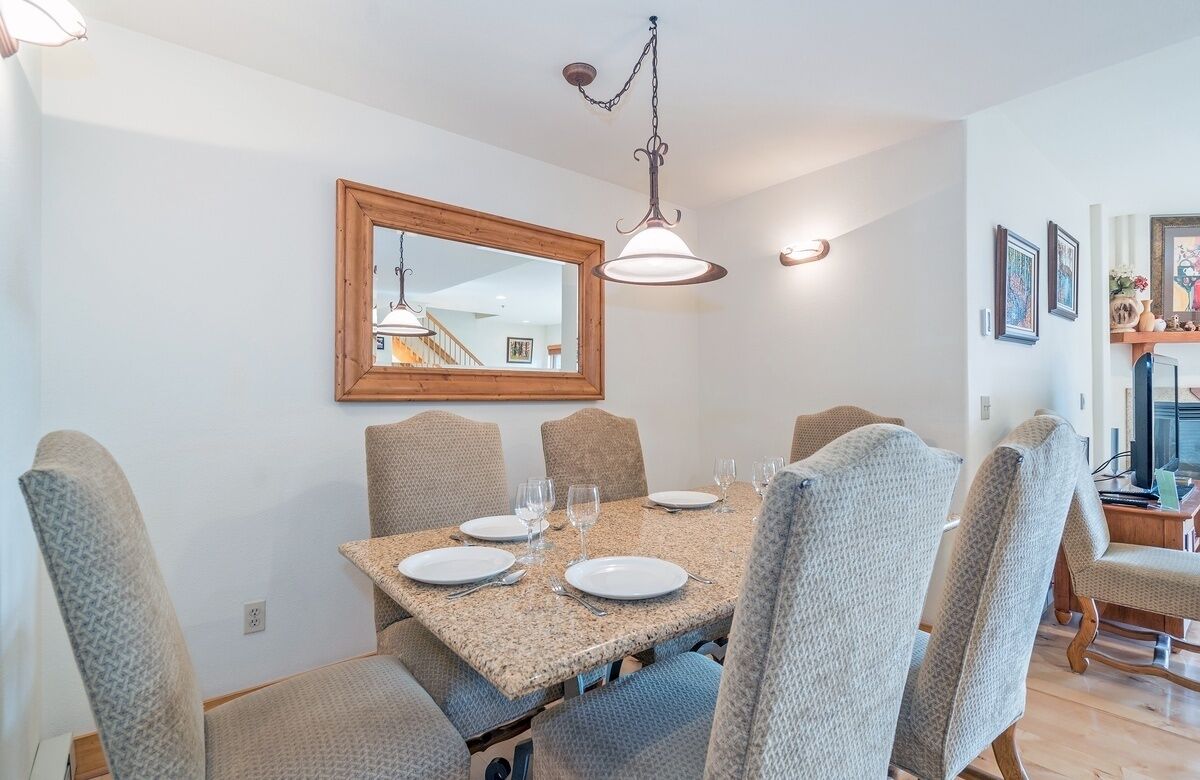 A dining area with a rectangular table set for six, surrounded by six upholstered chairs. A wooden-framed mirror hangs above the table, and a pendant light fixture is visible above.