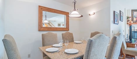 A dining area with a rectangular table set for six, surrounded by six upholstered chairs. A wooden-framed mirror hangs above the table, and a pendant light fixture is visible above.